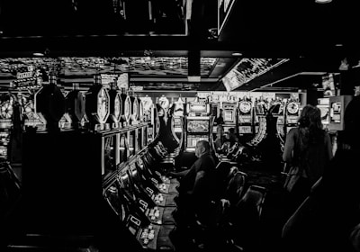 A dimly lit casino filled with rows of slot machines, some occupied by people. The machines' lights contrast with the dark surroundings, creating a vibrant yet subdued atmosphere.