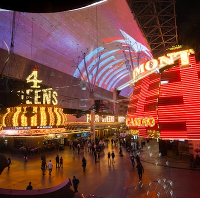 A bustling nighttime scene with illuminated casino signage towering over a busy pedestrian street. Neon lights in red and yellow highlight the names of various establishments. People are walking in different directions, enjoying the vibrant atmosphere.