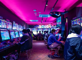 A vibrant arcade with rows of gaming machines adorned with colorful screens. Several people are immersed in playing games, seated on stools in a dimly lit room accented by an intense pink ceiling light. The atmosphere is lively and engaging.