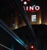 A night scene featuring a building with illuminated neon signage displaying 'CASINO'. The environment is urban with reflections on glass windows and glowing street traffic lights below. The atmosphere is dimly lit, with artificial lighting casting vibrant colors.